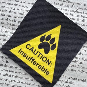 Könnte beinhalten: Ein quadratischer schwarzer Stoffaufnäher mit einem gelben Dreieck, einem schwarzen Pfotenabdruck-Symbol und dem Text CAUTION: Insufferable in schwarzen Großbuchstaben. Der Aufnäher hat eine gewebte Struktur und liegt auf einer bedruckten Seite.
