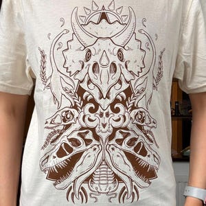 May include: A cream-colored t-shirt featuring a brown graphic print of multiple dinosaur skulls arranged in a symmetrical pattern. The design includes a large triceratops skull at the top, flanked by long-necked dinosaurs and smaller skulls.