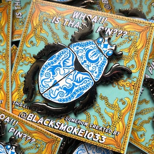 May include: A black and blue enamel pin featuring a stylised beetle with intricate designs. The pin has a gold border and the text "WHOA!! IS THAT F'N???" and "SOMEHOW CREATED BY @BLACKSMOKE1033".