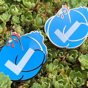May include: Two blue enamel pins shaped like chickens with white tick marks on them. The pins are on a bed of green succulents.
