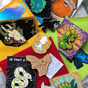 May include: An assortment of enamel pins and stickers with diverse designs, such as a snake, a dragon, a wolf, and a squid. The pins and stickers are displayed on colourful envelopes and cards. Some cards feature the text "WHOA!!! IS THAT A PIN???" and "THIS TOWN AIN'T BIG ENOUGH."