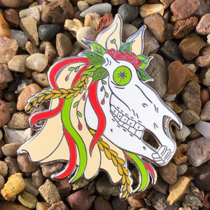 Op de afbeelding: Een witte emaille pin met een paarden schedel met een groen oog, omringd door rode, groene en gele bloemdetails. De pin ligt op een bed van kleine grijze stenen.