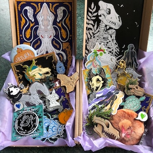 May include: Two open cardboard boxes filled with pins, stickers, and art prints. One box features a large print of a squid, while the other has a print of a skeletal animal. The boxes are lined with purple tissue paper, and the items are in a variety of colours.