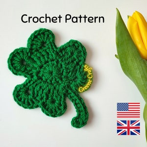 May include: A green crocheted four-leaf clover with a yellow "Greene Crafts" label. The clover is on a white background with a yellow tulip and flags of the United States and the United Kingdom.