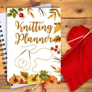 May include: A white notebook with a fall-themed cover that says "Knitting Planner". The cover features a watercolour illustration of leaves, pumpkins, and berries. A red knitted scarf and knitting needles are in the background.