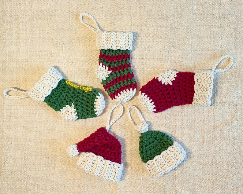 May include: Five miniature crocheted Christmas ornaments: two green and red stockings, two red and cream-colored hats, and one green and cream-colored hat. The stockings have cream-colored cuffs and feature a snowflake pattern. The hats have cream-colored brims. One stocking shows the text 'Greene Crafts' on its side. These charming handmade items are perfect for decorating a Christmas tree or adding a festive touch to gifts.