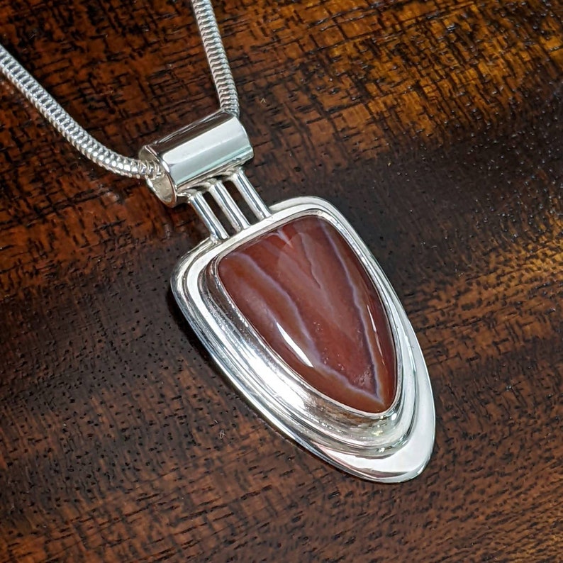 K&ouml;nnte beinhalten: Eine silberne Halskette mit einem Anh&auml;nger, der einen roten Achat-Stein in einem geometrischen Design tr&auml;gt.