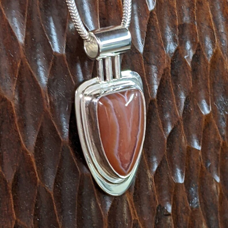 K&ouml;nnte beinhalten: Ein silberner Anh&auml;nger mit einem schildf&ouml;rmigen Design zeigt einen rot-wei&szlig;en Achat-Stein. Der Anh&auml;nger befindet sich an einer silbernen Kette.