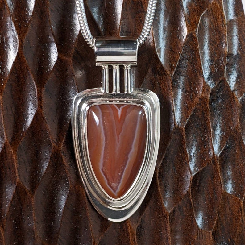 K&ouml;nnte beinhalten: Ein silberner Anh&auml;nger mit einem braunen und wei&szlig;en Achat-Stein, der in einem geometrischen Design gefasst ist. Der Anh&auml;nger h&auml;ngt an einer silbernen Kette.