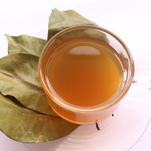 May include: A clear glass cup filled with a warm amber-colored liquid, resting on a matching glass saucer. Beside the cup, three dried, light brown, oval-shaped leaves are arranged, showing visible veins and a slightly curled, textured surface.