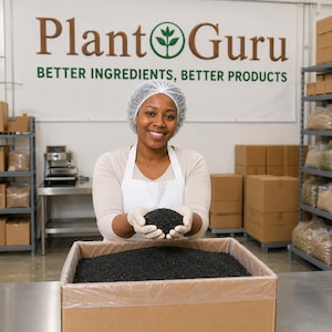 May include: A person wearing a white hairnet, white apron, and white gloves holds a handful of small black granules over a large cardboard box filled with the same black material. A banner behind reads Plant Guru BETTER INGREDIENTS, BETTER PRODUCTS.