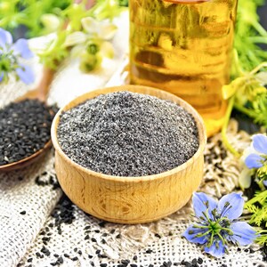 May include: A wooden bowl filled with gray seeds, a wooden spoon with black seeds, and a bottle of oil are displayed with blue flowers and green foliage. The seeds and oil are on a white cloth.