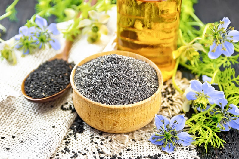 May include: A wooden bowl filled with gray seeds, a wooden spoon with black seeds, and a bottle of oil are displayed with blue flowers and green foliage. The seeds and oil are on a white cloth.