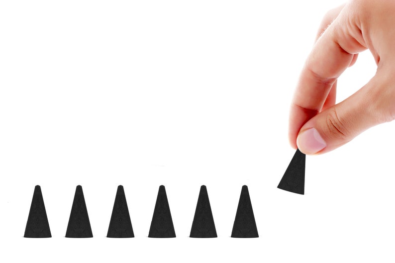 May include: A hand holding a black incense cone, with several other cones lined up on a white background. The cones are dark and have a triangular shape, suggesting a fragrance product.