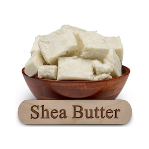 Może przedstawiać: Drewniana miska wypełniona kawałkami białego masła shea. Drewniany napis pod miską głosi "Shea Butter".