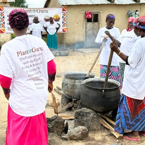 Puede incluir: Un grupo de personas con camisetas blancas con el texto "Plant Guru" y una lista de productos, reunidas alrededor de grandes ollas met&aacute;licas sobre un fuego. La escena tiene lugar al aire libre, cerca de un edificio con una pancarta.