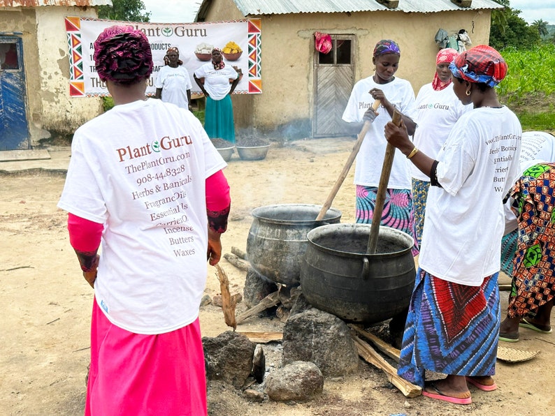 Puede incluir: Un grupo de personas con camisetas blancas con el texto "Plant Guru" y una lista de productos, reunidas alrededor de grandes ollas met&aacute;licas sobre un fuego. La escena tiene lugar al aire libre, cerca de un edificio con una pancarta.
