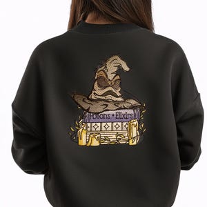 May include: Black sweatshirt featuring an embroidered design of a Sorting Hat atop books and candles. The books have the text "Potions + Elixirs". The design includes gold and tan colors, with a whimsical, magical theme.
