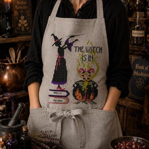 May include: A light gray linen apron featuring a cross-stitch design of a witch standing on books labeled Spells and Potions, next to a bubbling cauldron with legs sticking out and the text The Witch Is In. The apron has two front pockets and a tie.