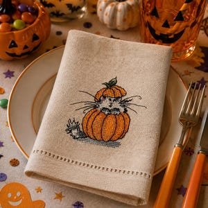 Könnte beinhalten: Eine beigefarbene Leinenserviette mit einem Kreuzstichmotiv, das eine kleine graue Katze zeigt, die aus einem leuchtend orangefarbenen Kürbis hervorschaut. Die Serviette hat einen dekorativen Hohlsaum am unteren Rand und liegt auf einem weißen Teller.