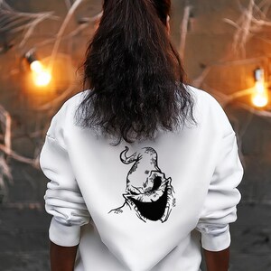 Może przedstawiać: Biała bluza z kapturem z czarną grafiką postaci z Miasteczka Halloween na plecach. Grafika to szczegółowy rysunek postaci w kształcie worka z uśmiechem. Bluza ma długie rękawy i kaptur.