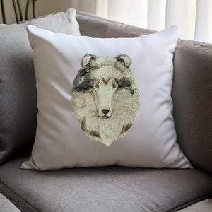 May include: White square throw pillow featuring an embroidered portrait of a Collie dog. The dog's face is detailed with shades of beige, gray, and black, set against a plain white background. The pillow is placed on a gray sofa.