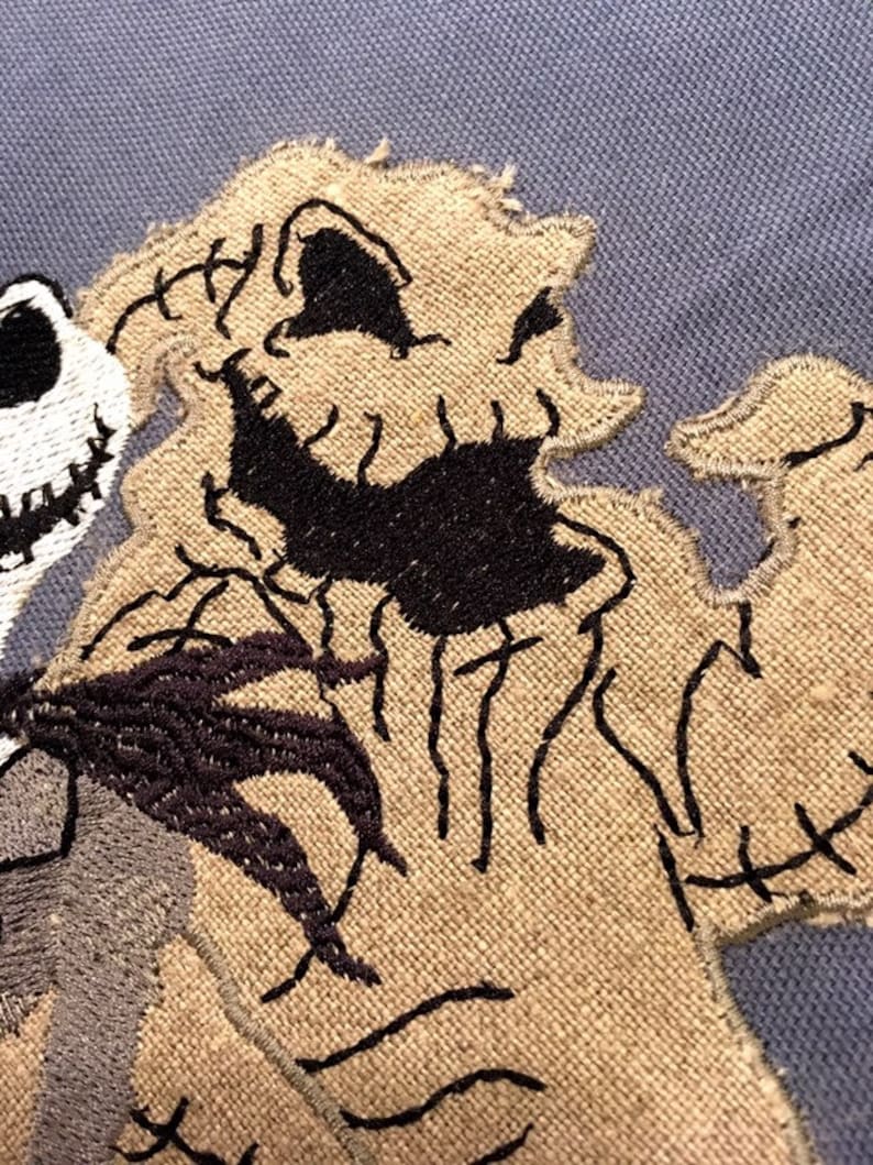 May include: Close-up of a brown and black embroidered patch featuring the character Jack Skellington from the movie The Nightmare Before Christmas. The patch is stitched with a detailed design of Jack's face and body.