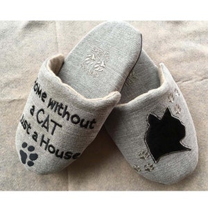 Pode incluir: Um par de pantufas de tecido cinza com pegadas bordadas e o texto "Home without a cat is just a house". Uma pantufa tem uma silhueta de gato preto.