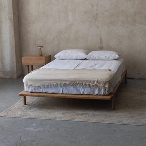 Peut inclure: Un lit en bois minimaliste avec une couverture de couleur claire et deux oreillers blancs. Une petite table de chevet en bois avec une lampe est à côté du lit. La pièce a une palette de couleurs neutres avec un tapis sur le sol.