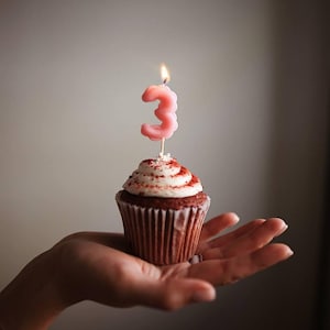 Könnte beinhalten: Ein Schokoladen-Cupcake mit weißem Zuckerguss und roten Streuseln, gekrönt von einer rosa Kerze mit der Zahl 3 und einer brennenden Flamme. Der Cupcake wird in einer Hand gehalten.