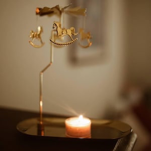 May include: A gold-colored candle carousel with rocking horse ornaments. The carousel is suspended above a small, round tray. A lit candle sits on the tray, casting a warm glow. The background is blurred, with soft lighting.