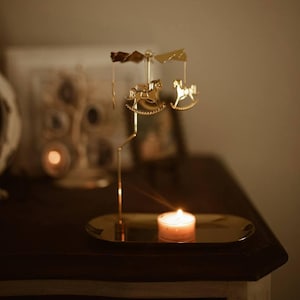 May include: A gold-colored candle holder with a rotating carousel design featuring rocking horse figures. The candle holder sits on a gold-colored oval tray, with a lit candle providing a warm glow. The background is blurred.