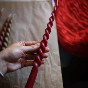 K&ouml;nnte beinhalten: Eine lange, schlanke, tiefrote Stabkerze mit einem durchgehenden Spiral- oder Drehdesign &uuml;ber die gesamte L&auml;nge. Die Kerze hat eine glatte, einfarbige Wachsoberfl&auml;che und einen einzelnen wei&szlig;en Docht am spitzen oberen Ende sowie einen geraden Fu&szlig;.
