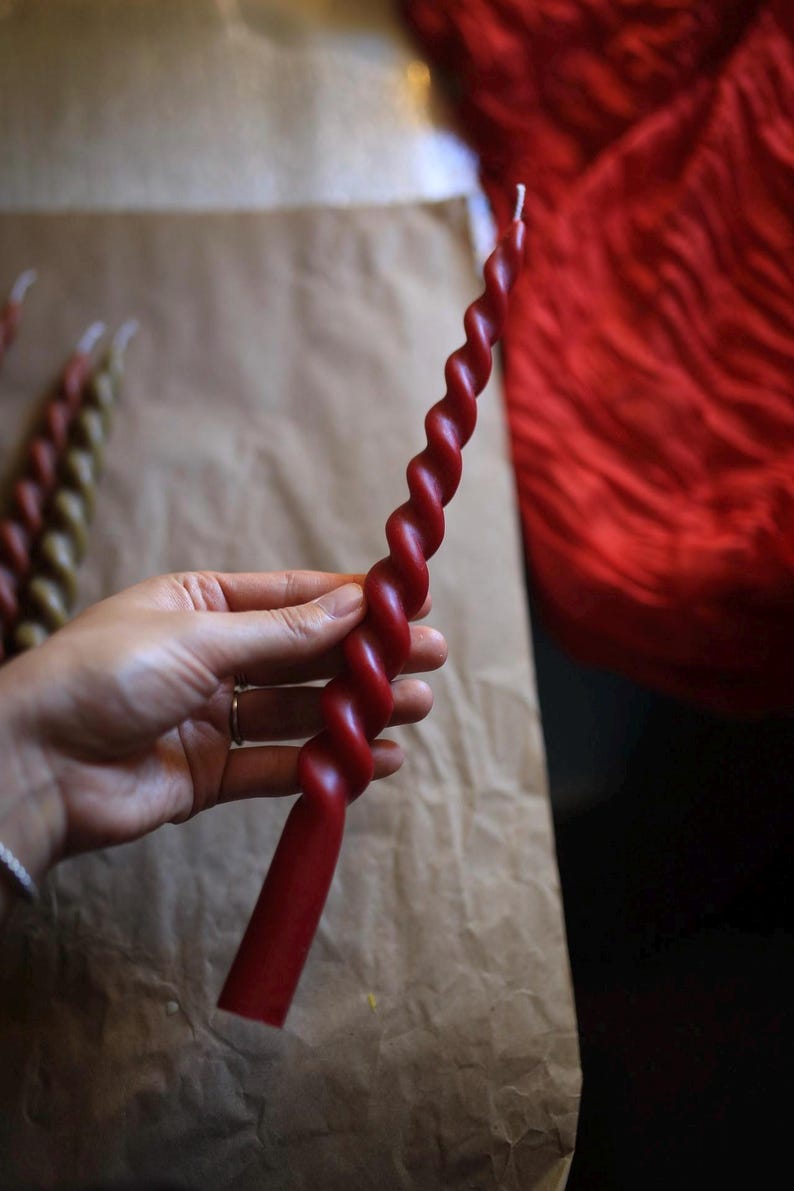 K&ouml;nnte beinhalten: Eine lange, schlanke, tiefrote Stabkerze mit einem durchgehenden Spiral- oder Drehdesign &uuml;ber die gesamte L&auml;nge. Die Kerze hat eine glatte, einfarbige Wachsoberfl&auml;che und einen einzelnen wei&szlig;en Docht am spitzen oberen Ende sowie einen geraden Fu&szlig;.