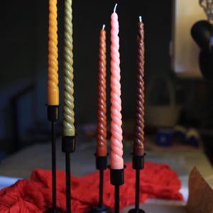 K&ouml;nnte beinhalten: F&uuml;nf hohe, schlanke, spiralf&ouml;rmige Stabkerzen in den Farben Gelb, Olivgr&uuml;n, Rostorange, Hellrosa und Dunkelbraun. Jede Kerze wird von einem schlichten schwarzen Kerzenst&auml;nder aus Metall mit rundem Fu&szlig; und d&uuml;nnem Schaft aufrecht gehalten.