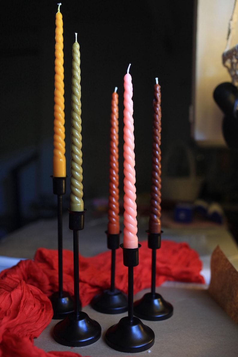 K&ouml;nnte beinhalten: F&uuml;nf hohe, schlanke, spiralf&ouml;rmige Stabkerzen in den Farben Gelb, Olivgr&uuml;n, Rostorange, Hellrosa und Dunkelbraun. Jede Kerze wird von einem schlichten schwarzen Kerzenst&auml;nder aus Metall mit rundem Fu&szlig; und d&uuml;nnem Schaft aufrecht gehalten.