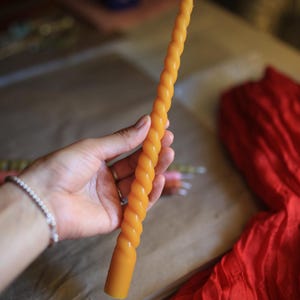 K&ouml;nnte beinhalten: Eine lange, schlanke, goldgelbe Stabkerze mit einem durchgehenden, gedrehten Spiralmuster &uuml;ber die gesamte L&auml;nge. Die Kerze hat eine glatte, wachsartige Textur und einen einzelnen wei&szlig;en Baumwolldocht am oberen Ende.
