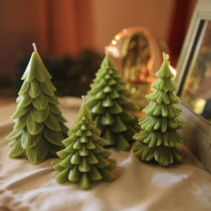 May include: Four green Christmas tree-shaped candles of varying sizes. The candles have detailed, textured designs mimicking the shape of a fir tree. The candles are arranged on a beige surface, suggesting a festive or holiday theme.