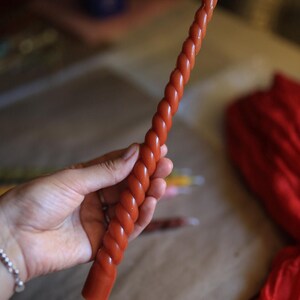 K&ouml;nnte beinhalten: Eine lange, schlanke, rostfarbene Stabkerze mit einem durchgehenden Spiral- oder gedrehten Design &uuml;ber die gesamte L&auml;nge. Die Kerze hat eine glatte Wachsoberfl&auml;che und einen kleinen, wei&szlig;en Baumwolldocht, der aus dem spitzen Ende herausragt.