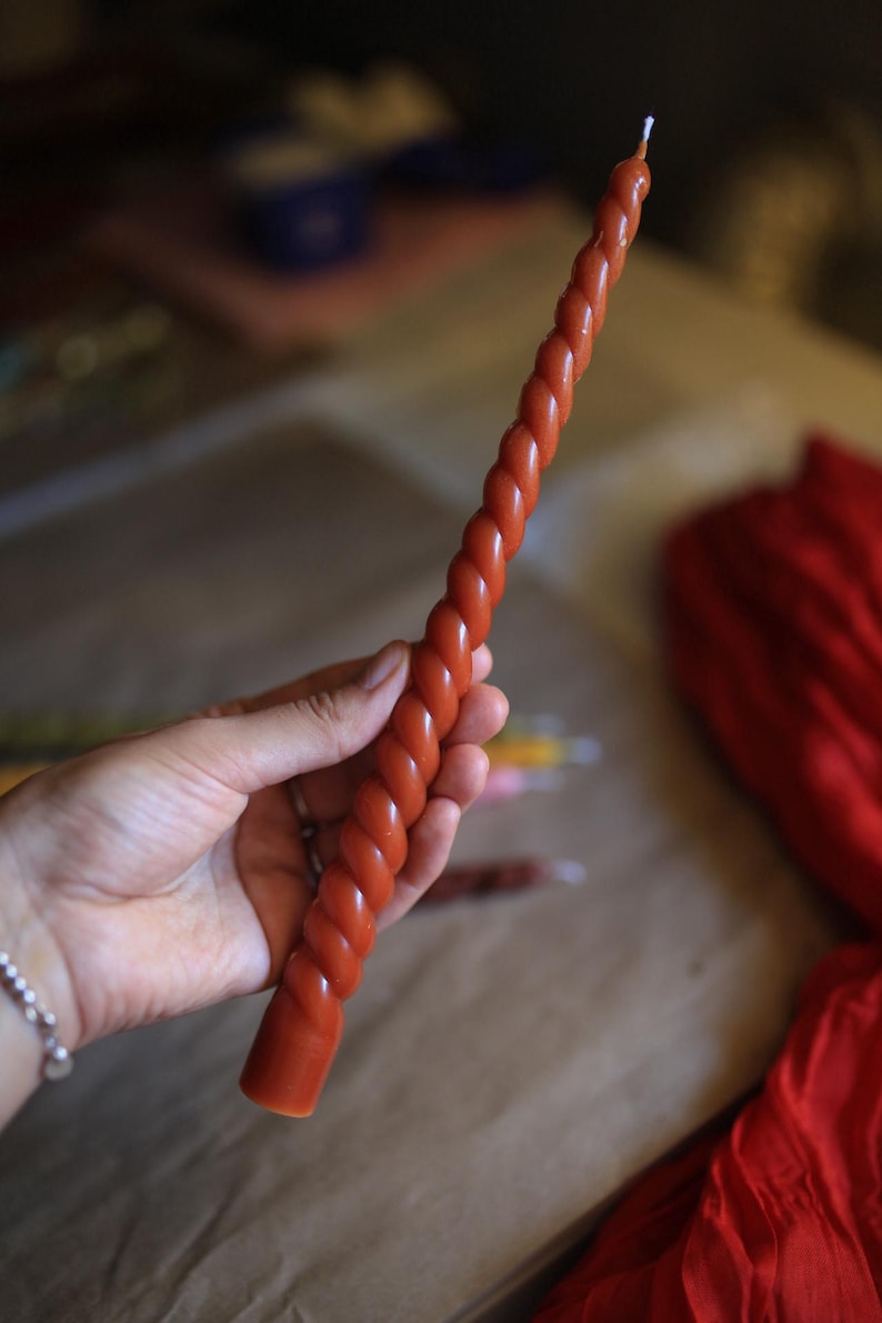 K&ouml;nnte beinhalten: Eine lange, schlanke, rostfarbene Stabkerze mit einem durchgehenden Spiral- oder gedrehten Design &uuml;ber die gesamte L&auml;nge. Die Kerze hat eine glatte Wachsoberfl&auml;che und einen kleinen, wei&szlig;en Baumwolldocht, der aus dem spitzen Ende herausragt.