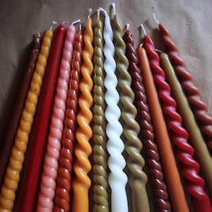 K&ouml;nnte beinhalten: Eine Sammlung von vierzehn Stabkerzen, die f&auml;cherf&ouml;rmig angeordnet sind. Das Set enth&auml;lt eine Mischung aus glatten und gedrehten Designs in verschiedenen Farben, darunter Braun-, Orange-, Rot-, Rosa-, Gelb-, Olivgr&uuml;n- und Wei&szlig;t&ouml;ne, jeweils mit Docht.
