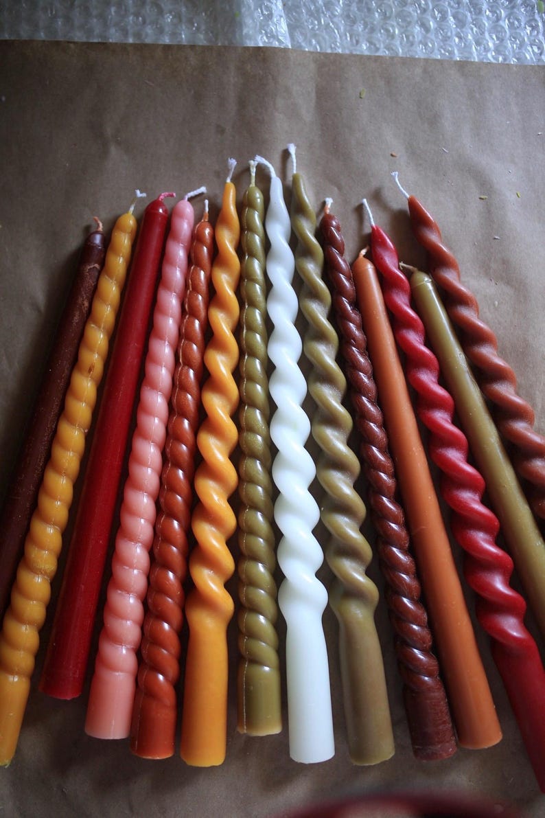 K&ouml;nnte beinhalten: Eine Sammlung von vierzehn Stabkerzen, die f&auml;cherf&ouml;rmig angeordnet sind. Das Set enth&auml;lt eine Mischung aus glatten und gedrehten Designs in verschiedenen Farben, darunter Braun-, Orange-, Rot-, Rosa-, Gelb-, Olivgr&uuml;n- und Wei&szlig;t&ouml;ne, jeweils mit Docht.