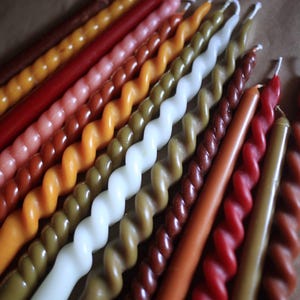 K&ouml;nnte beinhalten: Eine Sammlung langer, schlanker Stabkerzen, die in einer Reihe angeordnet sind. Die Kerzen weisen eine Mischung aus glatten und gedrehten Spiraldesigns in verschiedenen Erdt&ouml;nen auf, darunter Braun-, Rot-, Orange-, Gelb-, Olivgr&uuml;n- und Wei&szlig;t&ouml;ne.
