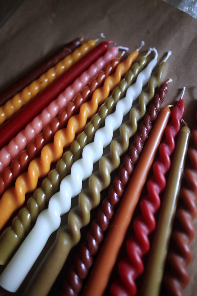 K&ouml;nnte beinhalten: Eine Sammlung langer, schlanker Stabkerzen, die in einer Reihe angeordnet sind. Die Kerzen weisen eine Mischung aus glatten und gedrehten Spiraldesigns in verschiedenen Erdt&ouml;nen auf, darunter Braun-, Rot-, Orange-, Gelb-, Olivgr&uuml;n- und Wei&szlig;t&ouml;ne.
