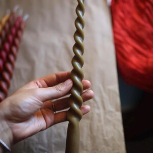 K&ouml;nnte beinhalten: Eine lange, olivgr&uuml;ne Stabkerze mit einem markanten, spiralf&ouml;rmig gedrehten Design &uuml;ber die gesamte L&auml;nge. Die Kerze hat eine glatte Wachsoberfl&auml;che und einen einzelnen wei&szlig;en Docht an der Spitze. Sie ist am Fu&szlig; zylindrisch und verj&uuml;ngt sich nach oben.