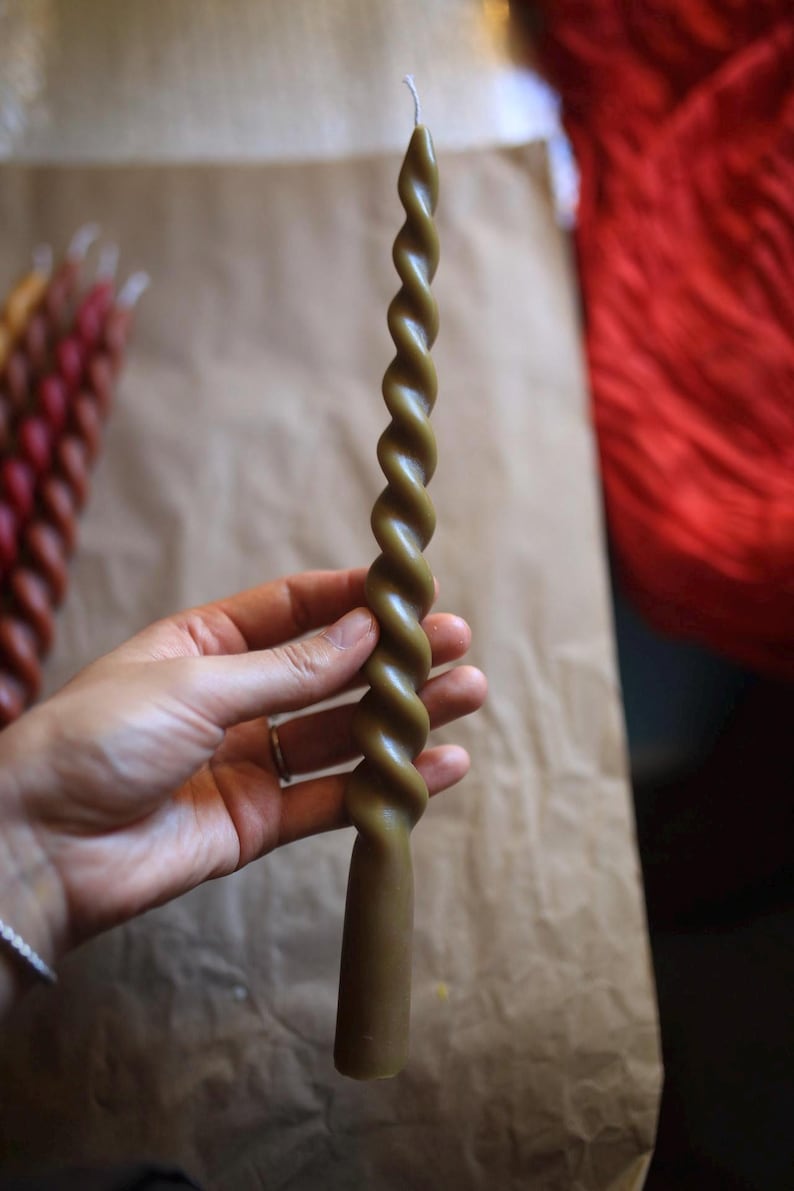 K&ouml;nnte beinhalten: Eine lange, olivgr&uuml;ne Stabkerze mit einem markanten, spiralf&ouml;rmig gedrehten Design &uuml;ber die gesamte L&auml;nge. Die Kerze hat eine glatte Wachsoberfl&auml;che und einen einzelnen wei&szlig;en Docht an der Spitze. Sie ist am Fu&szlig; zylindrisch und verj&uuml;ngt sich nach oben.