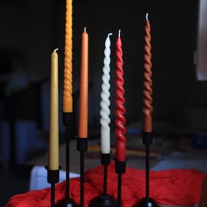 K&ouml;nnte beinhalten: Eine Sammlung von sechs Stabkerzen in verschiedenen Gelb-, Orange-, Rot- und Wei&szlig;t&ouml;nen, die sowohl glatte als auch gedrehte Spiraldesigns aufweisen. Jede Kerze wird von einem schlanken schwarzen Metallkerzenhalter mit rundem Fu&szlig; gehalten.