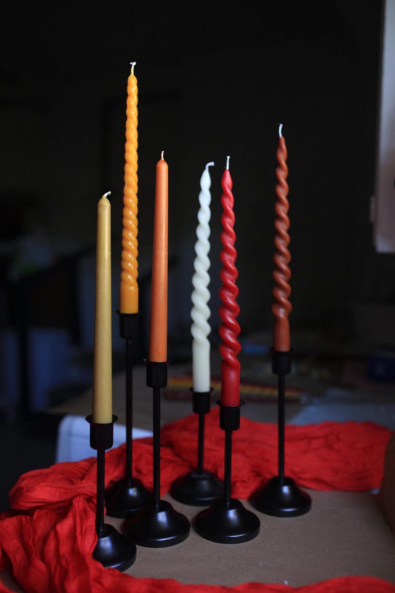 K&ouml;nnte beinhalten: Eine Sammlung von sechs Stabkerzen in verschiedenen Gelb-, Orange-, Rot- und Wei&szlig;t&ouml;nen, die sowohl glatte als auch gedrehte Spiraldesigns aufweisen. Jede Kerze wird von einem schlanken schwarzen Metallkerzenhalter mit rundem Fu&szlig; gehalten.