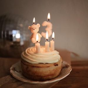 May include: A small cake with white frosting, topped with four lit candles. One candle is shaped like a teddy bear, and another forms the number 3. The cake sits on a decorative plate, creating a festive birthday scene.