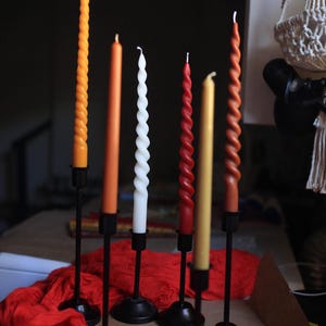 K&ouml;nnte beinhalten: Sechs Stabkerzen in verschiedenen Farben und Texturen, darunter gedrehte und glatte Designs in Orange, Wei&szlig;, Rot und Gelb, die jeweils von einem schlanken schwarzen Metallkerzenhalter mit rundem Fu&szlig; aufrecht gehalten und gruppiert sind.
